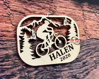 May include: A wooden ornament featuring a silhouette of a person riding a mountain bike through a mountain landscape. The ornament is personalized with the name "HALEN" and the year "2020".