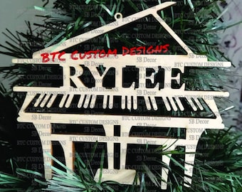 May include: A wooden Christmas ornament shaped like a piano with the name "RYLEE" written on it in red letters. The ornament is decorated with the words "BTC Custom Designs" and "SB Decor" repeated throughout.