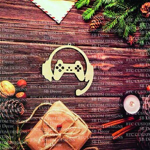 May include: A wooden ornament shaped like a video game controller with headphones. The ornament is on a wooden background with a gift box, pine cones, and cinnamon sticks.