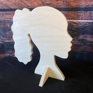 May include: A wooden silhouette of a person's profile, featuring a ponytail. The cutout is light-colored and stands upright with a small wooden stand. The background is a dark surface with a wooden wall.
