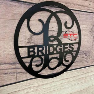 May include: Black metal circle with the letters "B" and "C" intertwined in a decorative font. The name "BRIDGES" is written in a straight font below the intertwined letters.