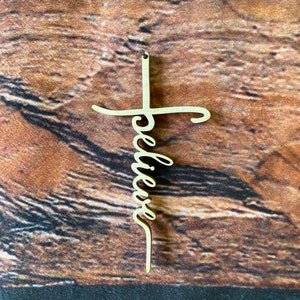 K&ouml;nnte beinhalten: Ein Holzkreuz mit dem Wort "glauben" in Kursivschrift darauf. Das Kreuz ist hellbraun und h&auml;ngt an einem Holzgrund.