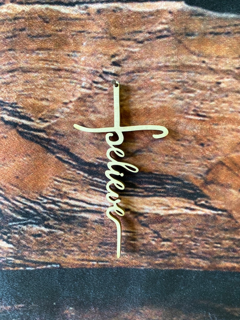 K&ouml;nnte beinhalten: Ein Holzkreuz mit dem Wort "glauben" in Kursivschrift darauf. Das Kreuz ist hellbraun und h&auml;ngt an einem Holzgrund.
