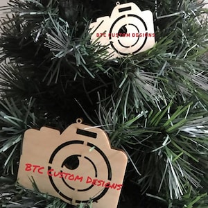 May include: Two wooden camera-shaped Christmas ornaments hanging on a green Christmas tree. The ornaments are light brown with a cutout lens design and the text "BTC CUSTOM DESIGNS" in red.