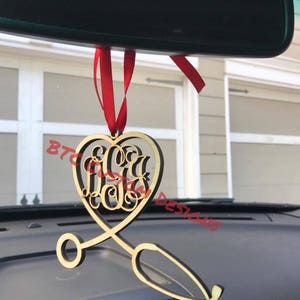 May include: A wooden heart-shaped ornament with a stethoscope attached. The heart has a monogram with the letters "JCU" inside. The ornament is hanging from a red ribbon in a car.