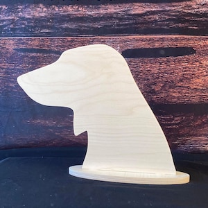 May include: A wooden cutout of a dog's head, standing on a round base. The cutout is unpainted and made of light wood.