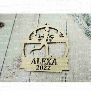 Peut inclure: Une d&eacute;coration de No&euml;l en bois repr&eacute;sentant une silhouette de coureur avec des arbres en arri&egrave;re-plan. La d&eacute;coration est personnalis&eacute;e avec le nom "ALEXA" et l'ann&eacute;e "2022".
