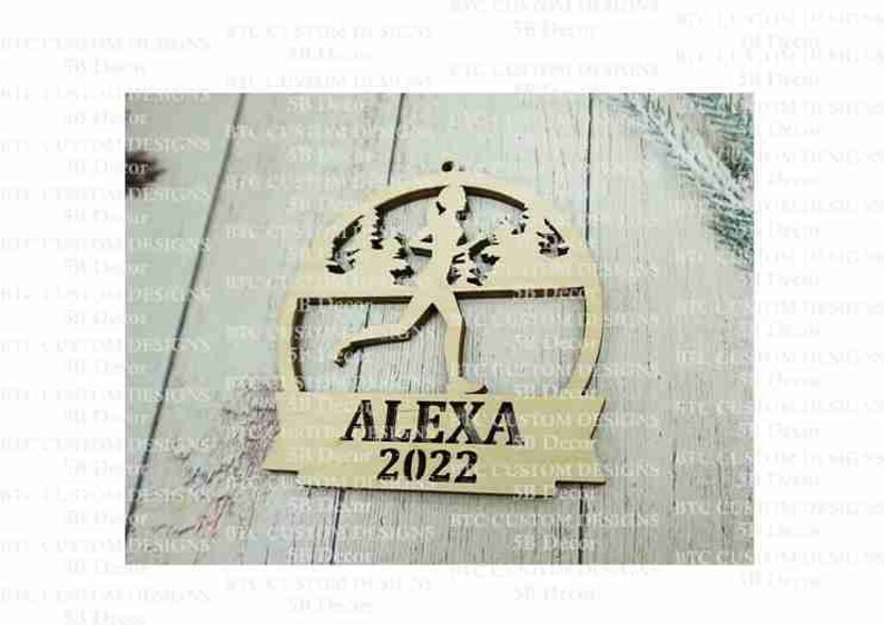 Peut inclure: Une d&eacute;coration de No&euml;l en bois repr&eacute;sentant une silhouette de coureur avec des arbres en arri&egrave;re-plan. La d&eacute;coration est personnalis&eacute;e avec le nom "ALEXA" et l'ann&eacute;e "2022".