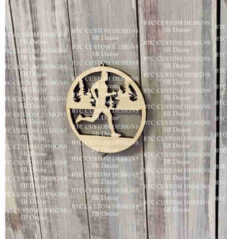 Peut inclure: Une d&eacute;coration en bois repr&eacute;sentant la silhouette d'un coureur dans un d&eacute;cor de for&ecirc;t. La d&eacute;coration est circulaire et poss&egrave;de un trou en haut pour l'accrocher.