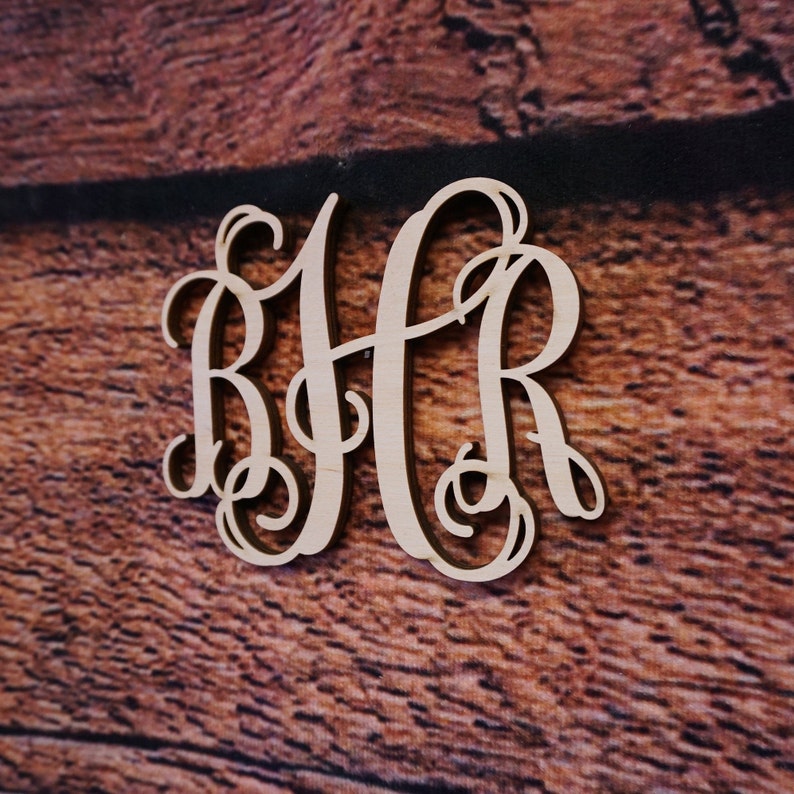 May include: A wooden monogram with the letters "BHR" in a cursive script font. The letters are cut out of a light brown wood and are arranged in a traditional monogram style.