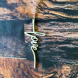 May include: A wooden cross with the word "love" written on it in a cursive font. The cross is a light brown color and is hanging from a string.