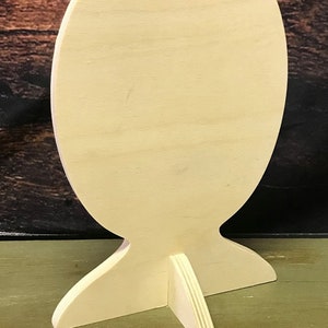 May include: A blank, oval-shaped wooden sign with a stand. The sign is made of light-colored wood and has a smooth surface. The stand is made of three pieces of wood that are joined together to form a triangular base.
