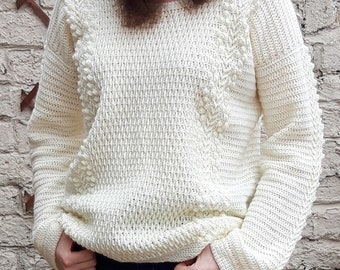 May include: A white crocheted sweater with a textured stitch pattern and a scalloped neckline. The sweater has a relaxed fit and long sleeves.