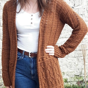 May include: A brown knitted cardigan with a cable knit pattern. The cardigan is open at the front and has long sleeves. The person is wearing a white shirt and blue jeans.