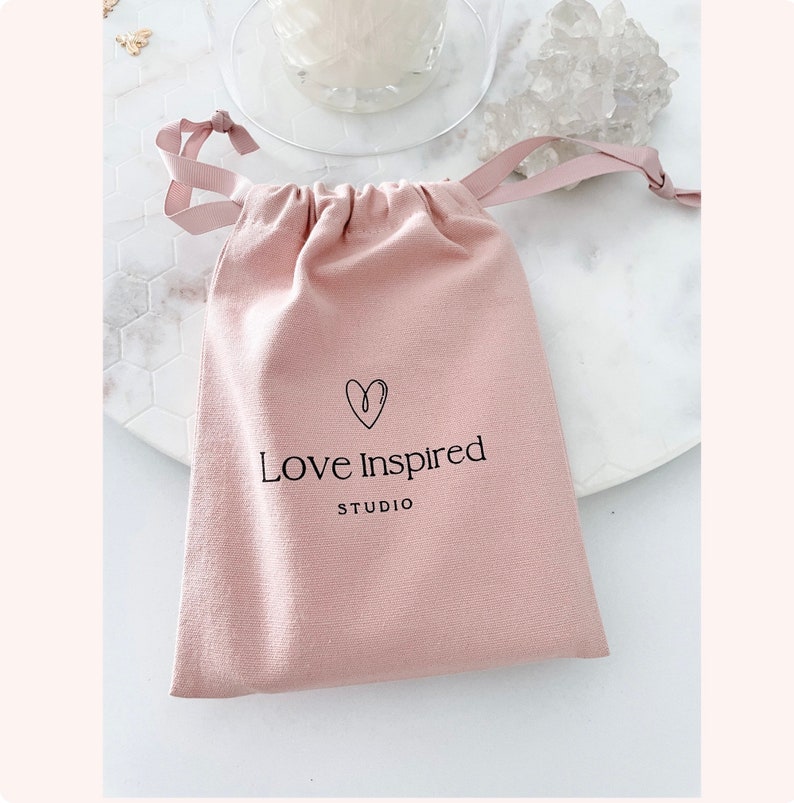 Puede incluir: Una bolsa de tela rosa con cord&oacute;n de ajuste con un coraz&oacute;n y el texto "Love Inspired Studio" impreso en la parte delantera.