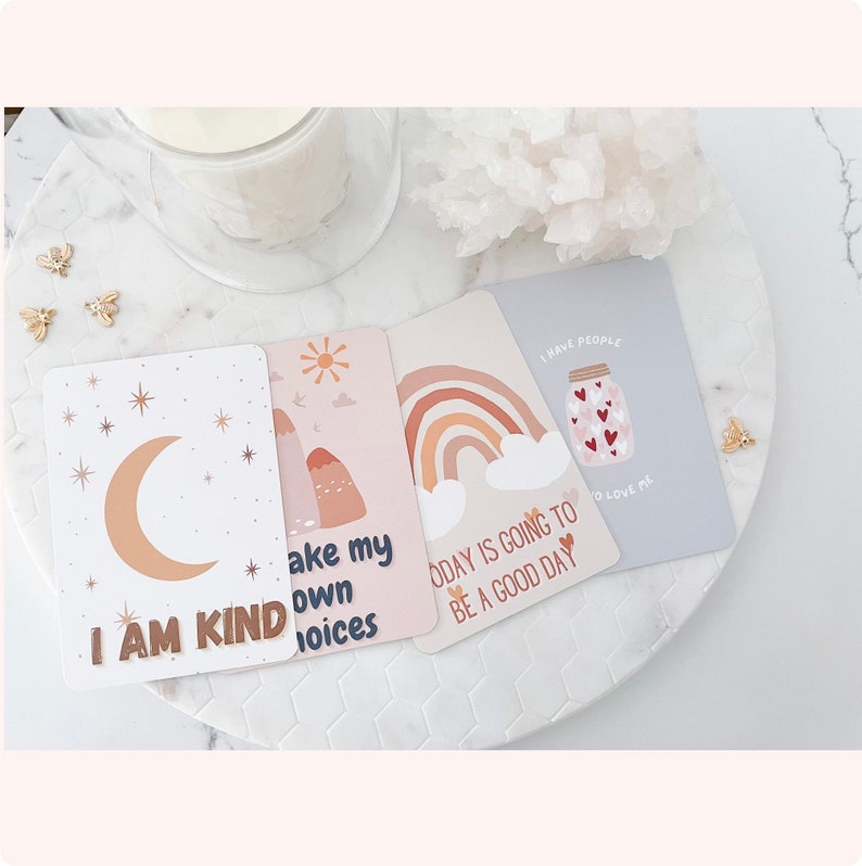 Puede incluir: Cuatro tarjetas de afirmaci&oacute;n con mensajes positivos. Las tarjetas son rosas y blancas con ilustraciones de una luna, monta&ntilde;as, un arco&iacute;ris y un frasco lleno de corazones. El texto en las tarjetas dice "Soy amable", "Tomo mis propias decisiones", "Hoy va a ser un buen d&iacute;a", y "Tengo gente que me quiere".