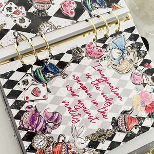 May include: A black and white checkered planner with a whimsical Alice in Wonderland theme. The planner features illustrations of Alice, the Mad Hatter, the Cheshire Cat, and other characters from the story. The text "Imagination is the only weapon in the war against reality" is printed on the cover.