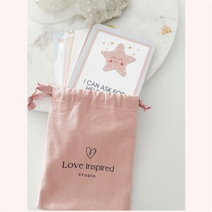 Puede incluir: Una bolsa de cord&oacute;n rosa con el texto "Love Inspired Studio" y un dise&ntilde;o de coraz&oacute;n. La bolsa contiene un juego de tarjetas con ilustraciones de una estrella de mar sonriente y el texto "Puedo pedir ayuda".