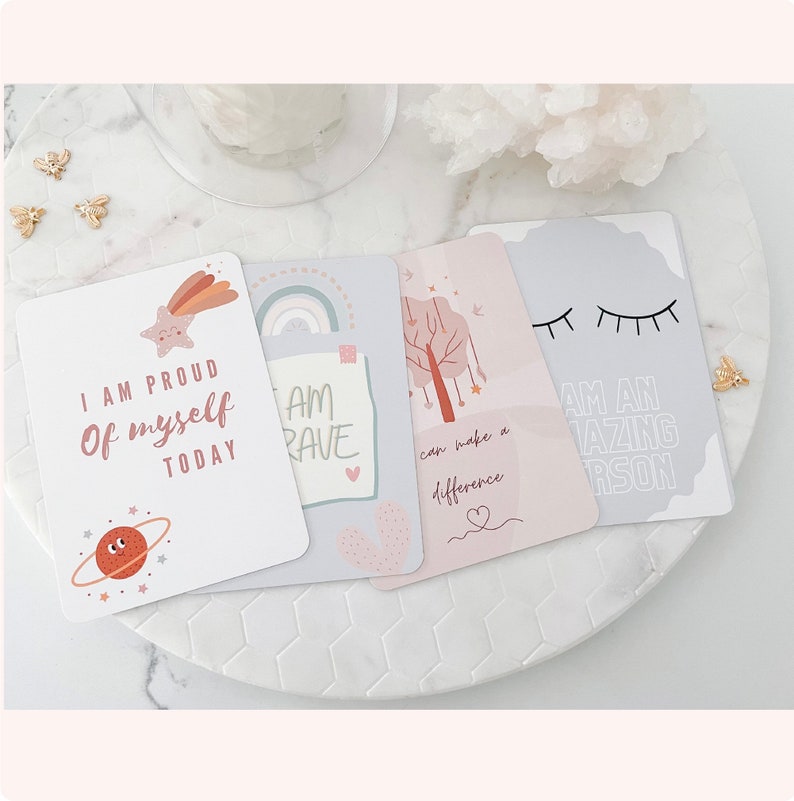 Puede incluir: Cinco tarjetas de afirmaci&oacute;n con mensajes positivos, que incluyen "Estoy orgulloso de m&iacute; mismo hoy", "Soy valiente", "Puedo marcar la diferencia" y "Soy una persona incre&iacute;ble". Las tarjetas est&aacute;n decoradas con ilustraciones de una estrella, un arco&iacute;ris, un &aacute;rbol y pesta&ntilde;as.