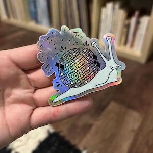 Puede incluir: Una pegatina holográfica de un caracol que lleva una bola de discoteca con notas musicales a su alrededor.
