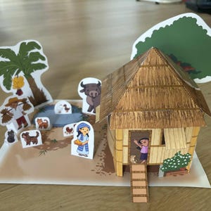 May include: A papercraft diorama depicting a rural scene with a thatched-roof house, palm tree, and various animal and human figures. The scene includes a pond and a person waving from the doorway.