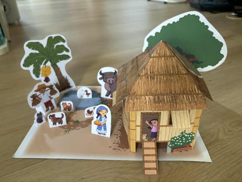 May include: A papercraft diorama depicting a rural scene with a thatched-roof house, palm tree, and various animal and human figures. The scene includes a pond and a person waving from the doorway.