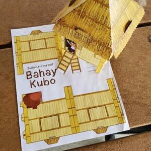 May include: A completed paper model of a Bahay Kubo, a traditional Filipino house, with a thatched roof and bamboo-style walls. The model is placed on top of the printed instructions. The text "Build-It-Yourself Bahay Kubo" is visible.