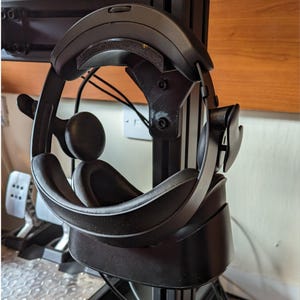 May include: A black VR headset is hanging on a stand. The headset has a black headband and a black faceplate. The headset is hanging on a black stand with a silver knob.