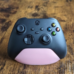 May include: A black Xbox controller with a pink base. The controller features a black and white directional pad, black buttons, and a black and white Xbox logo.