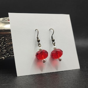 Peut inclure: Une paire de boucles d'oreilles à perles rouges facettées avec des crochets d'oreilles en bronze antique et de petits accents de perles blanches. Les boucles d'oreilles sont présentées sur une carte blanche.