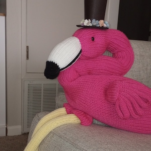 May include: A pink knitted flamingo with yellow legs wearing a black top hat with a floral trim. The flamingo is sitting on a gray couch.