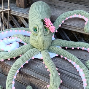 May include: A green knitted octopus with pink pom-poms around the tentacles and a pink flower on its head.