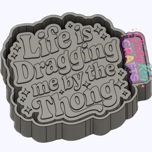 May include: A grey cookie cutter featuring the text "Life is Dragging me by the Thong" in a retro font. The cutter has a cloud-like shape with star accents and a rectangular handle with the "Sticky Key Crafts" logo.