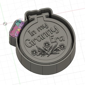 May include: A gray, 3D-printed cookie cutter in the shape of an embroidery hoop. The text "In my Granny Era" is embossed on the inside, along with a floral design. A small tag with text is attached to the side.