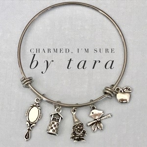 May include: Silver-toned charm bracelet with multiple charms, including a mirror, tower, gnome, scarecrow, and apple. The text "CHARMED, I'M SURE" and "by tara" are displayed on the image.