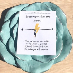 May include: A white card with the text "Be stronger than the storm." and a yellow lightning bolt graphic. The card also includes instructions for a wish bracelet: "Close your eyes and make a wish tie this bracelet on your wrist. When the bracelet breaks in two, this is when your wish comes true."