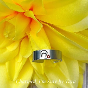May include: A silver ring with a small red heart and a white outline of a ghost on it. The ring is on a yellow flower.