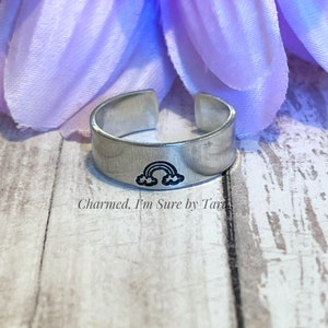 May include: A silver adjustable ring with a rainbow and clouds design. The ring is engraved with the text "Charmed. I'm Sure by Tara".