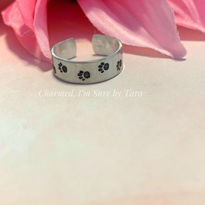 May include: A silver-colored adjustable ring with black paw prints stamped around the band. The ring is displayed on a light pink surface with a pink flower in the background. The text "Charmed. I'm Sure by Tara" is visible.