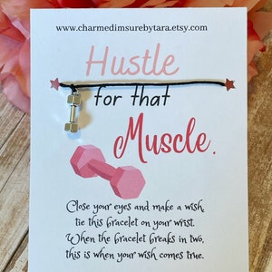 May include: A white card with black text that reads "Hustle for that Muscle."  A pink dumbbell charm is attached to a black string bracelet. The card also includes instructions for making a wish with the bracelet.