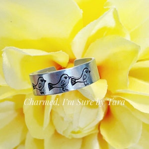 May include: A silver adjustable ring with three small birds etched into the band. The ring is on a yellow flower.