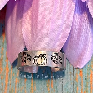 May include: A silver ring with a pumpkin and leaves design. The ring is engraved with the text "Charmed, I'm Sure by Tara".