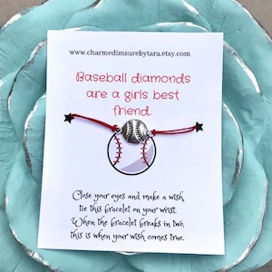 May include: A white card with red text that reads "Baseball diamonds are a girls best friend."  A red string bracelet with a silver baseball charm is attached to the card. The card also includes the text "Close your eyes and make a wish, tie this bracelet on your wrist. When the bracelet breaks in two, this is when your wish comes true."