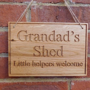 Puede incluir: Un letrero de madera con el texto "Grandad's Shed Little helpers welcome" tallado en él. El letrero está colgado en una pared de ladrillo.