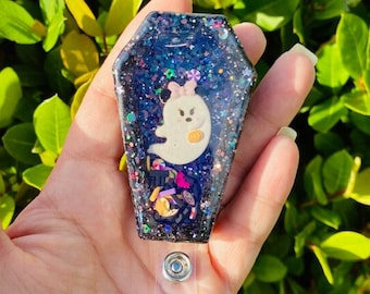 May include: Black glitter resin coffin-shaped badge holder with a white ghost and candy inside. The holder is attached to a clear retractable badge reel.