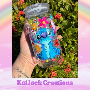May include: A clear glass jar with a lid, featuring a hand-painted image of the character Stitch from Lilo & Stitch. The jar is decorated with colorful flowers. The text "KatJack Creations" and Etsy shop URL are visible.