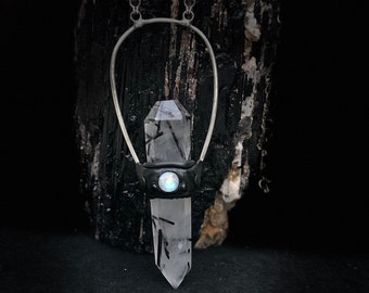 May include: A silver chain necklace with a black leather and silver cage pendant featuring a clear quartz crystal with black inclusions and a moonstone accent.