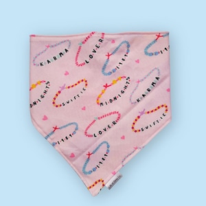 Puede incluir: Bandana rosa con un patrón repetido de diseños de pulseras coloridas. Cada pulsera tiene una palabra como "Karma", "Lover" y "Swiftie". Pequeños corazones rosas están dispersos. Hecho de tela suave.
