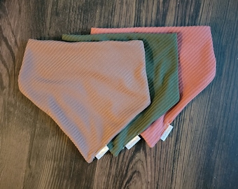 May include: Three fabric dog bandanas in pink, green, and brown. The bandanas are folded and stacked on top of each other. The bandanas have a ribbed texture.