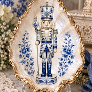 Peut inclure: Une figurine de casse-noisette décorative ornée de motifs floraux bleus et blancs, placée dans un grand plat en forme de coquillage irisé avec un bord doré texturé. Le casse-noisette tient un long sceptre et porte un uniforme assorti.